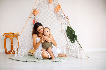 Young happy family loving mother with baby kid sitting together in wigwam teepee at home, mom...