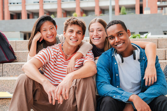 Portrait of a group of real multiracial high school students smiling and looking at camera sitting on the steps of a university campus stairway. International teenage friends laughing. Young people - Powered by Adobe