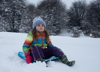 Fototapeta na wymiar Little girl with rainbow jacket sledging down a snowy hill. High quality photo