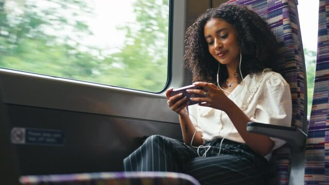 Young businesswoman commuting sitting by window on moving train streaming film or show to mobile phone wearing earphones - shot in slow motion