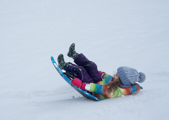 Fototapeta na wymiar Little girl with rainbow jacket sledging down a snowy hill, falling in a snow. High quality photo