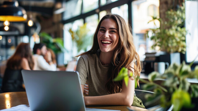Young woman seated and smiling at the camera, with a laptop in front of her and a blurred office environment in the background