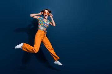 Full length photo of lovely young lady jumping running touch sunglass wear trendy flared pants isolated on dark blue color background