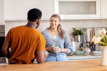 Smiling young mixed couple standing in kitchen drinking coffee at home.
