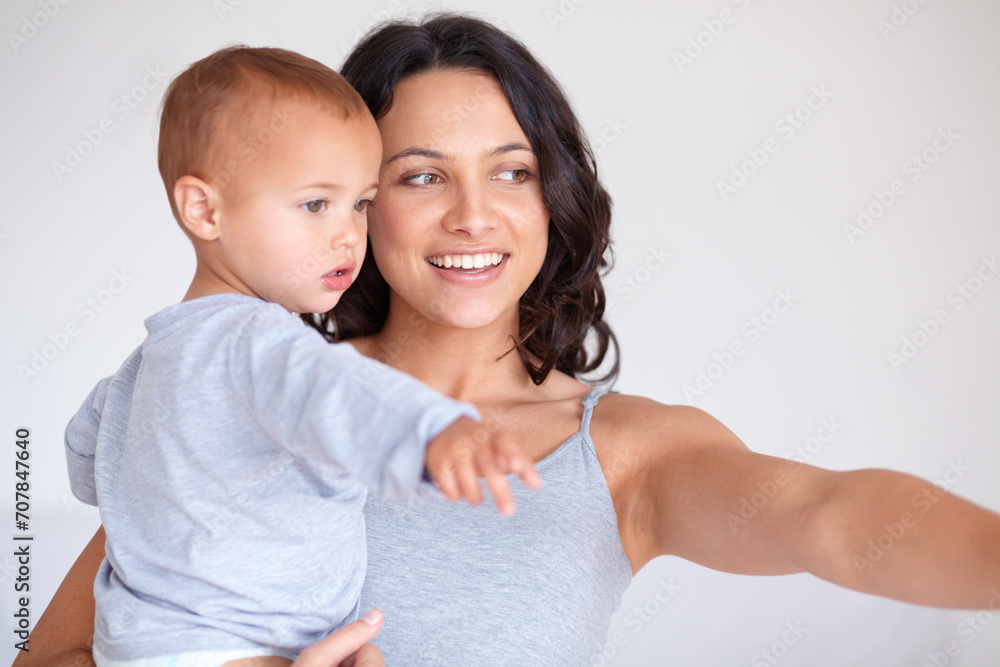 Wall mural Mom holding toddler with smile, pointing and child care with support at home in morning. Happy woman, baby boy and bonding together in bedroom with development, trust and safety with mothers love.