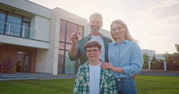 Happy family of three standing outside near big house. Smiling family of four at background of yard. Father holding keys of new house. Young family buying new appartment. Satisfied parents and kid.