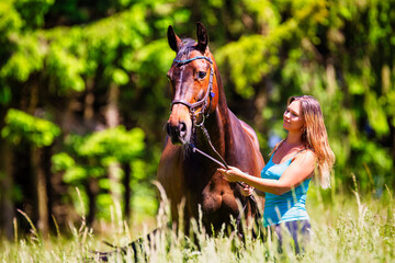 A young woman rider with long brunette hair stands with her horse on a high summer meadow in the...