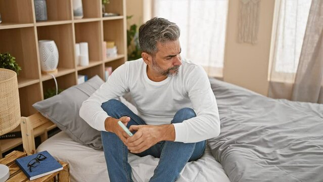 Stressed young hispanic grey-haired man sitting in bedroom, texting message on smartphone, worried expression