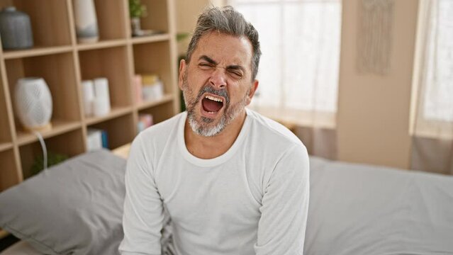 Exhausted young hispanic man with grey hair, yawning while relaxing on comfortable bed in his bedroom, lifestyle expression captured indoors