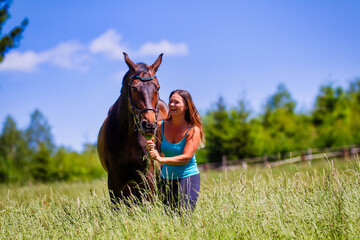 A young woman rider with long brunette hair stands with her horse on a high summer meadow in the...