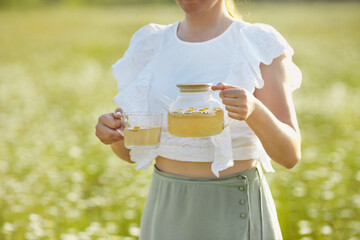 Woman  holding glass transparent teapot and mug with chamomile tea, standing in camomile field....