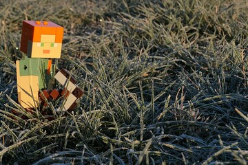 Fototapeta premium LEGO Minecraft figure of Alex with iron pickaxe walking through winter frozen grass, morning sunshine. 