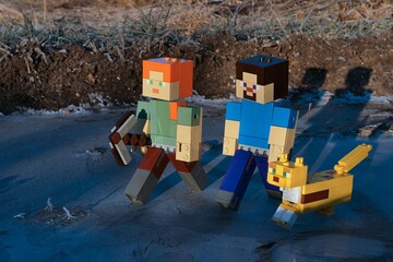 Fototapeta premium LEGO Minecraft figures of Alex with iron pickaxe, smiling Steve and their pet friend Ocelot walking on icy surface in winter morning sunshine. 
