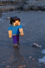 Fototapeta premium LEGO Minecraft figure of smiling Steve walking on ice frosty surface near asphalt road, sunlit by early morning winter sunshine.
