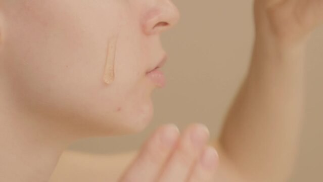 Extreme close-up shot of face Caucasian young woman puts skin care serum on her cheek from dropper on beige background. Skin care serum advertising, copy space.