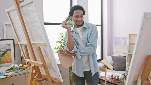 A thoughtful man with curly hair holds a paintbrush and palette in a bright art studio.