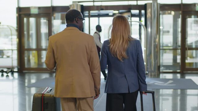 Back view of male and female business partners walking with suitcases to exit door and speaking while leaving hotel, bellhop passing by with luggage cart
