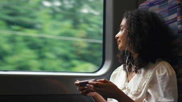 Young businesswoman commuting sitting by window on moving train streaming film or show to mobile phone wearing earphones looking out of window - shot in slow motion