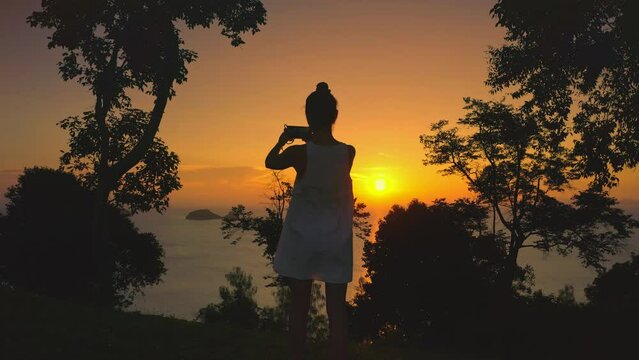 Woman make sunset panorama enjoy sea sunset view. Woman silhouette standing in forest take photo on mobile phone. Bright orange sky sun in background. Exotic nature landscape. Travel, holiday relax