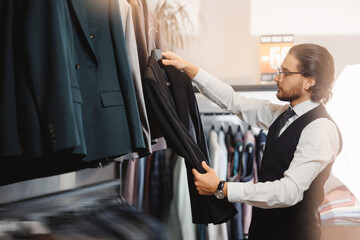 Store classic clothes for men. Smiling businessman in glasses chooses to buy suit jacket in shop.