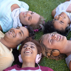 Friends, headphones and group relax on grass at park, listening to music or radio in summer. Audio, smile and students on lawn in top view, nature and young people streaming sound together outdoor