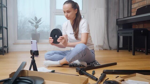 A vlogger girl shoots DIY video content with an overview of parts for self-assembly of furniture on a phone camera mounted on a tripod for subscribers of her channel.