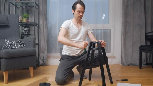 A man repairs a chair on his own, kneeling in front of hand tools and furniture parts laid out on cardboard, and tightening the screws with a ratchet screwdriver.