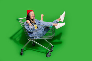 Full size photo of pretty young girl raise fists enjoy ride shopping cart dressed stylish denim outfit isolated on green color background