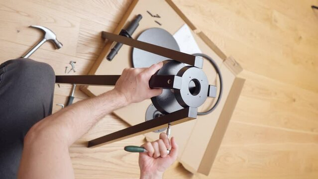 A man, using hand tools, a hammer and a ratchet screwdriver, manually screws the screws into the legs of a chair that he unpacked from a box and assembles himself at home. Vertical shot.