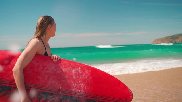 Young woman tourist carrying red surfboard walking towards sea. Female in bikini enjoying surfing on summer vacation. Healthy active lifestyle.