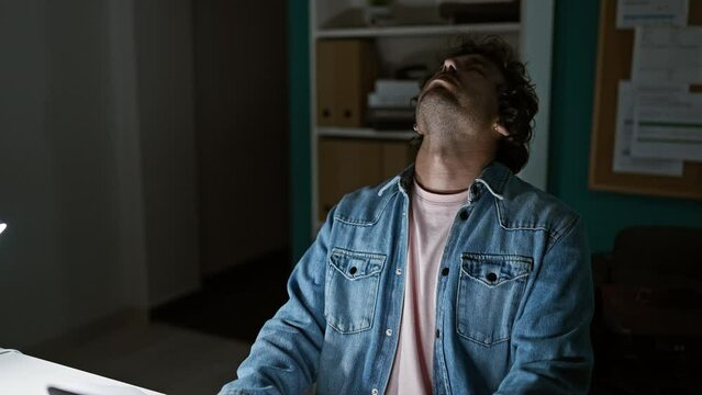 A tired hispanic man in a denim jacket expressing frustration in a dark office.