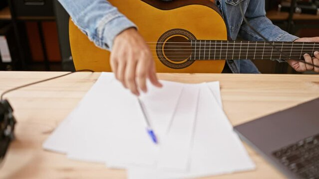 Focused man composing music while playing guitar indoors, with notation sheets and laptop on wooden table.