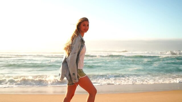 Portrait of smiling blond woman standing arms outstretched on sandy beach. Beautiful female in denim jacket and shorts standing near big rock and sea waves. Lady enjoys summer vacation.