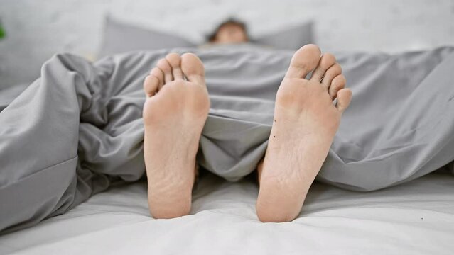 A person relaxing in bed with bare feet protruding from under a gray comforter in a bright bedroom setting.
