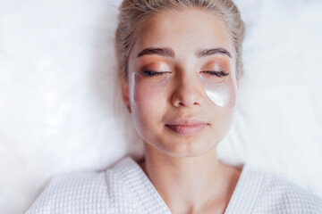 Beautiful blonde woman in bathrobe is lying with silicone patches under her eyes in spa salon or...
