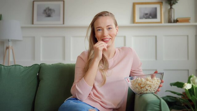 young woman with blonde hair sits on couch in living room and watch movie on TV eating popcorn. comedy funny serial sets laughing and smiling. looks pleased and happy