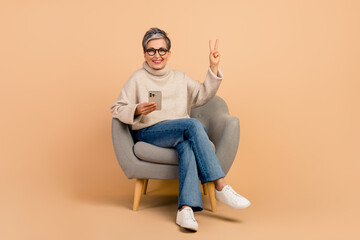 Full body size photo of senior attractive woman sitting armchair showing v sign using new iphone...