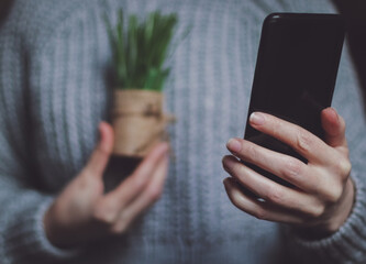 The hand of a Caucasian young girl holds a black smartphone in front of her and shows a flower in a pot in it.