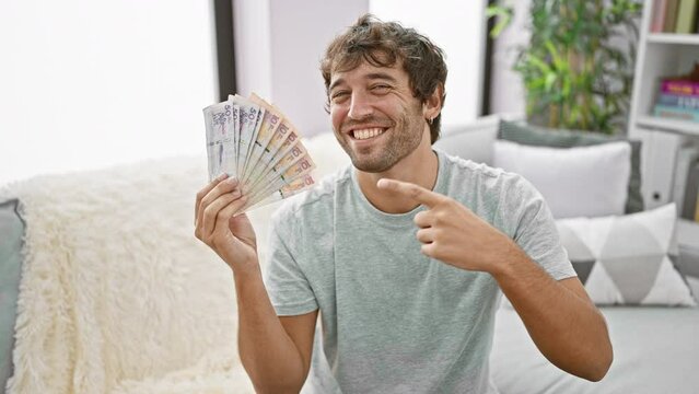Beaming young man, holding colombian pesos at home, confidently pointing and smiling with joy