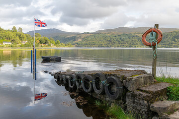 Union Jack flag on a pole with a boat tied up at high tide in sea Loch Sunart at Strontian,...