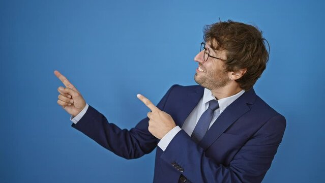 Cheery young man dons business suit, flashing a dazzling smile looking at the camera. with both hands pointing to the side, over isolated blue wall, welcoming you in!