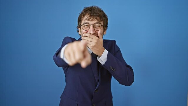 Cheerful young man in business suit gives a joyous laugh, pointing and pranking you with a smirk on his isolated blue background, hand over mouth!