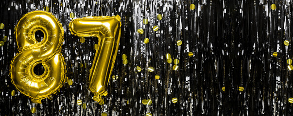 Gold foil balloon number number 87 on a background of black tinsel decoration. Birthday greeting...