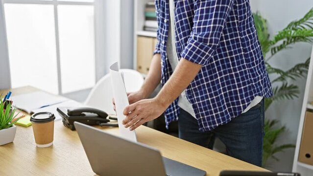 Smiling young business worker, a handsome blond, standing firm in his office, juggling work documents with online tasks on his laptop