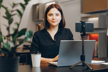 Young woman participates in online video conference through laptop, workplace in cafe. Phone on...