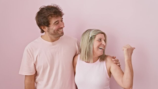 Happy caucasian mother and son joyfully pointing thumb up together, standing with open mouth smiles over isolated pink background
