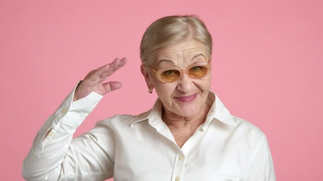 Old blonde woman with short hair light make-up standing over pink background giving respect salute with a smile on her face, laughing heartily afterwards. High quality 4k footage