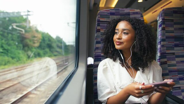 Front view of young businesswoman commuting sitting by window on moving train streaming film or show to mobile phone wearing earphones looking out of window - shot in slow motion