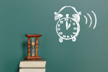 A vintage hourglass standing on a book against the backdrop of a chalkboard with an alarm clock drawn in chalk. There is copy space on the board for inserting text or drawing - 707980472