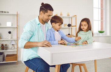 Father playing with his two kids boy and a girl in a puzzle on the living room at home. Young smiling man collecting jigsaw with his children on the table. Family and people concept.